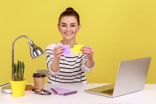 Happy smiling woman putting together two colored pieces of jigsaw, easily solving difficult tasks at work, sitting at workplace with laptop. Indoor studio studio shot isolated on yellow background.