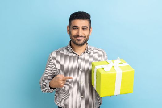 Happy businessman with beard standing, holding yellow gift box, pointing finger, looking at camera with toothy smile, wearing striped shirt. Indoor studio shot isolated on blue background.