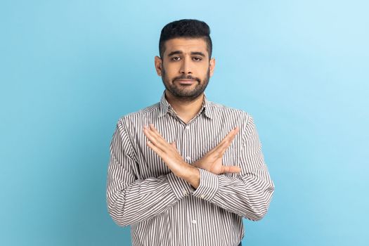 There is no way. Concerned businessman crossing hands showing x gesture, rejecting bullying and abuse seriously looking at camera, wearing striped shirt. Indoor studio shot isolated on blue background
