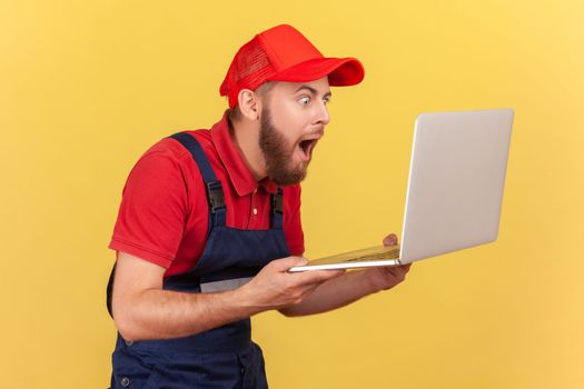 Side view of extremely shocked handyman standing laptop in hands and looking at notebook display with big eyes, wearing uniform and red cap. Indoor studio shot isolated on yellow background.
