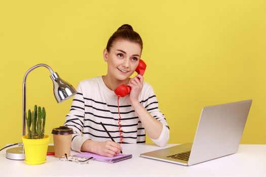 Portrait of smiling woman talking on red retro phone, looking away with dreamy facial expression, sitting at workplace and writing in notepad. Indoor studio studio shot isolated on yellow background.