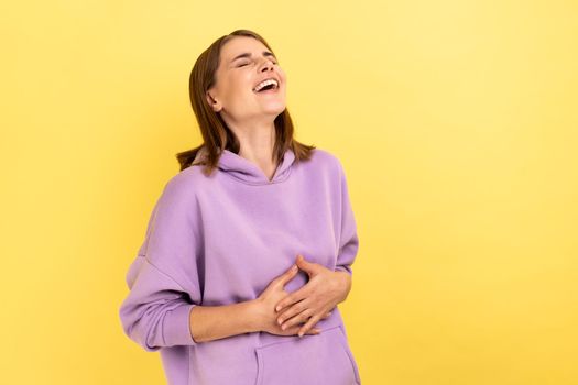 Portrait of happy woman with dark hair holding belly, laughing, hearing funny joke or anecdote, being in good mood, wearing purple hoodie. Indoor studio shot isolated on yellow background.