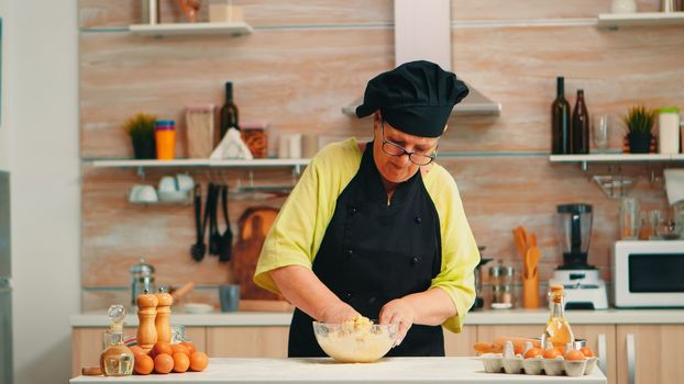 Woman wearing chef bonete while mixing cracked eggs with flour in kitchen while preparing food following traditional recipe. Retired elderly baker kneading in glass bowl ingredients for homemade cake