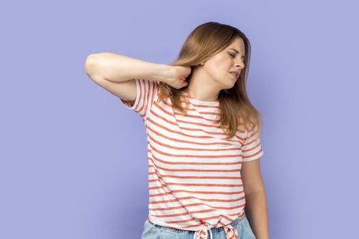 Portrait of attractive tired upset unhealthy blond woman wearing striped T-shirt standing massaging neck to relieve pain, muscle strain in back. Indoor studio shot isolated on purple background.