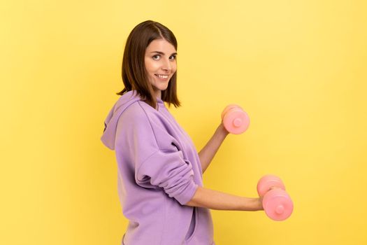 Side view of woman standing doing fitness exercises with pink dumbbells, training hands, looking at camera with smile, wearing purple hoodie. Indoor studio shot isolated on yellow background.