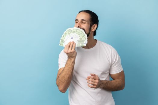 Portrait of avaricious rich man with beard wearing white T-shirt smelling earned euro banknotes, enjoying success and big profit, wealthy life. Indoor studio shot isolated on blue background.