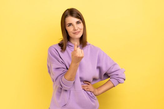 Come here. Portrait of attractive woman making beckoning gesture, inviting to come, flirting, looking at camera, wearing purple hoodie. Indoor studio shot isolated on yellow background.