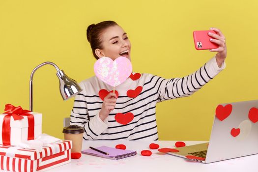 Attractive happy woman manager sitting at workplace with laptop and holding heart on sticks, making selfie or having video call. Indoor studio studio shot isolated on yellow background.