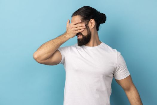 Don't want to look at this. Portrait of scared man with beard wearing white T-shirt standing, covering eyes with hand, refusing to watch. Indoor studio shot isolated on blue background.