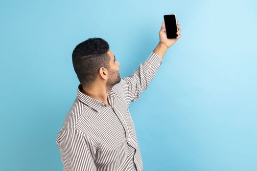 Back view of businessman talking video call, taking selfie on mobile phone, holding smartphone up with mockup blank display, wearing striped shirt. Indoor studio shot isolated on blue background.