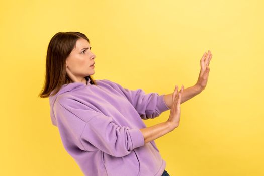 No, it's scary. Side view of frightened shocked woman raising hands in fear, looking horrified and panicking, hiding from phobia wearing purple hoodie. Indoor studio shot isolated on yellow background