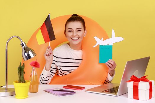 Portrait of delighted woman office manager with rubber ring holding passport and German flag, being happy to travel to Germany. Indoor studio studio shot isolated on yellow background.