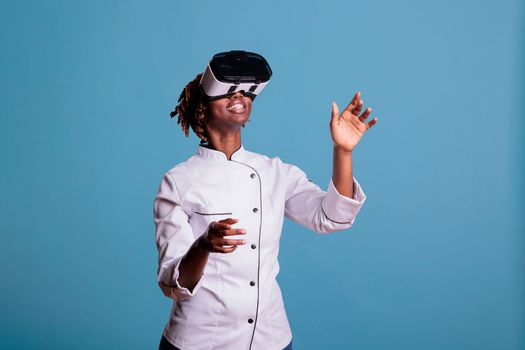 Professional female chef in uniform playing virtual reality games with VR goggles, exploring simulations. African american cook moving arms while touching objects in cyberspace.