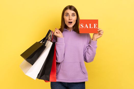 Extremely surprised woman holding in hands paper shopping bags and sale card, looking at camera with big eyes and opened mouth, wearing purple hoodie. Indoor studio shot isolated on yellow background.
