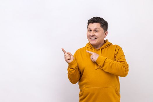 Look at advertise. Positive man pointing finger away at empty space for advertisement, looking at camera with toothy smile, wearing urban style hoodie. Indoor studio shot isolated on white background.