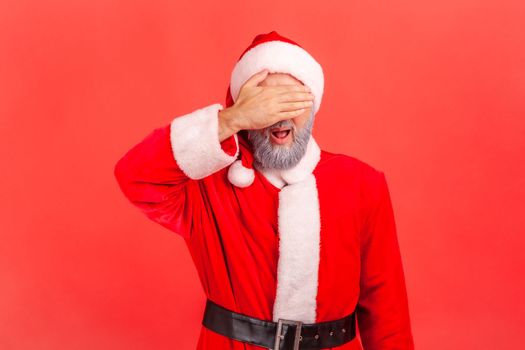 Portrait of elderly man with gray beard wearing santa claus costume covering eyes with palm, does not want to see, avoiding problems. Indoor studio shot isolated on red background.
