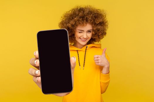 Portrait of joyful pleased woman with Afro hairstyle wearing casual style hoodie showing thumb up and big smart phone empty screen, recommend new app. Indoor studio shot isolated on yellow background.