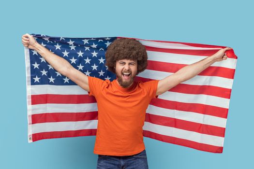 Portrait of man with Afro hairstyle in T-shirt holding USA flag and looking at camera with rejoicing look, screaming, celebrating national holiday. Indoor studio shot isolated on blue background.