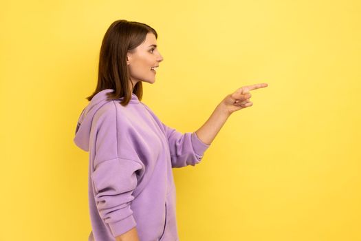 Side view of optimistic beautiful young woman standing and pointing at copyspace with happy positive face, wearing purple hoodie. Indoor studio shot isolated on yellow background.