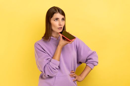Portrait of woman talking to smartphone using digital voice app, recording message, searching information on internet, wearing purple hoodie. Indoor studio shot isolated on yellow background.
