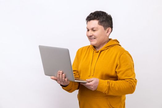 Attractive middle aged man working on laptop typing email or chatting in social network, happy face expression, wearing urban style hoodie. Indoor studio shot isolated on white background.
