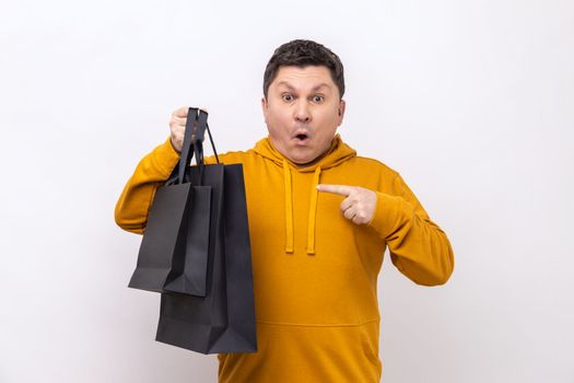Astonished middle aged man pointing finger at black paper bags with purchases, shocked with seasonal sale, wearing urban style hoodie. Indoor studio shot isolated on white background.