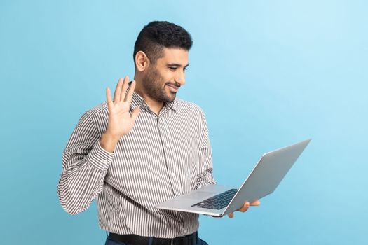 Businessman waving hand gesturing hello to laptop screen, talking on video call with coworker, communicating online conference, wearing striped shirt. Indoor studio shot isolated on blue background.