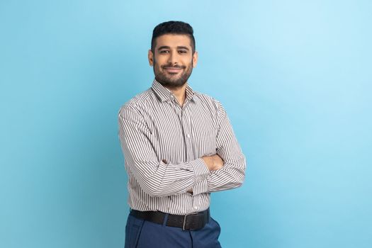 Young happy successful businessman posing with happy confident expression, looking at camera, keeping arms folded, wearing striped shirt. Indoor studio shot isolated on blue background.