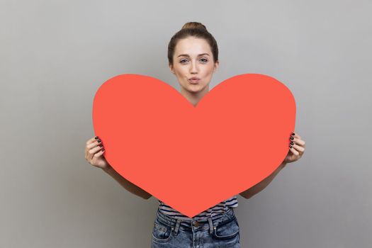 Portrait of romantic pretty woman wearing striped T-shirt holding big red heart and sending air kissing, looking at camera. Indoor studio shot isolated on gray background.