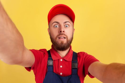 Surprised amazed worker man with wearing red cap and blue uniform taking selfie, looking at camera with big eyes POV, point of view of photo. Indoor studio shot isolated on yellow background.