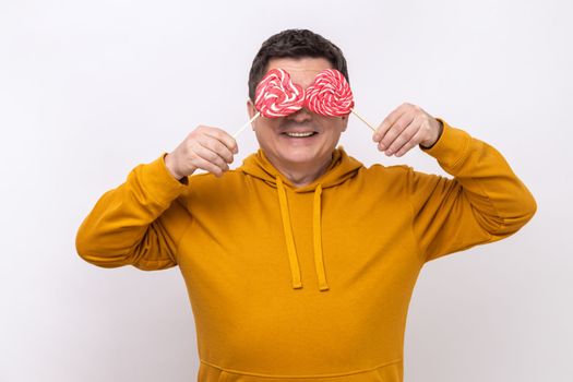 Portrait of funny man covering eyes with sweet heart shaped candy lollipop, showing tasty sugary dessert, wearing urban style hoodie. Indoor studio shot isolated on white background.