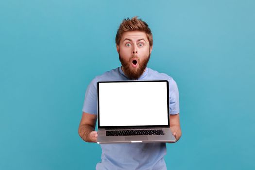 Portrait of bearded man holding and showing laptop with blank empty display, place for your advertisement, looking at camera with big eyes and open mouth Indoor studio shot isolated on blue background