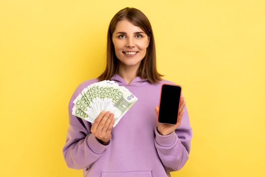 Positive satisfied dark haired woman holding euro banknotes and cell phone with empty screen, online betting, winning, wearing purple hoodie. Indoor studio shot isolated on yellow background.