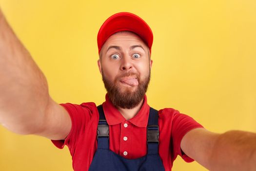 Childish worker man wearing red cap and blue uniform taking selfie, looking at camera and showing tongue out POV, point of view of photo. Indoor studio shot isolated on yellow background.