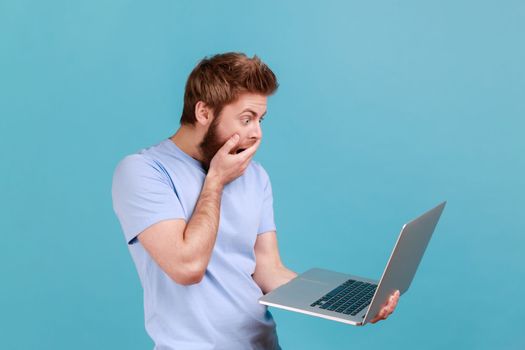 Portrait of handsome astonished surprised bearded man looking at laptop display with big eyes shocked with content, covered mouth with palm. Indoor studio shot isolated on blue background.
