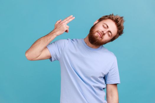 Portrait of tired depressed bearded man holding fingers near temple pretending holding gun, going to make shot, faced with problems and crisis. Indoor studio shot isolated on blue background.