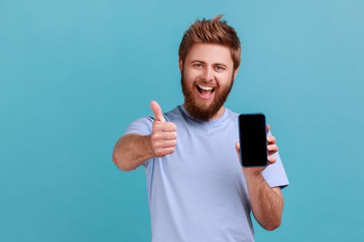 Portrait of positive optimistic bearded man standing holding phone with empty display, showing thumbs up with toothy smile, looking at camera. Indoor studio shot isolated on blue background.