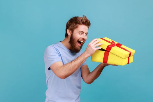 Portrait of excited handsome bearded man unboxing yellow gift box, looking inside with positive facial expression, birthday present. Indoor studio shot isolated on blue background.