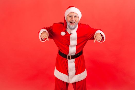 Happy excited elderly man with gray beard wearing santa claus costume pointing down with fingers showing place for idea presentation, commercial text. Indoor studio shot isolated on red background.