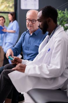 African american medic consulting senior patient with glucometer instrument while measuring insulin and glucose level from blood sample. Elderly man having checkup visit consultation in hospital lobby