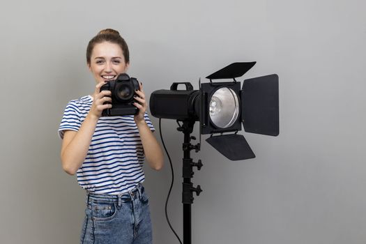 Portrait of smiling satisfied woman photographer wearing striped T-shirt standing with photocamera and reflector, looking at camera. Indoor studio shot isolated on gray background.