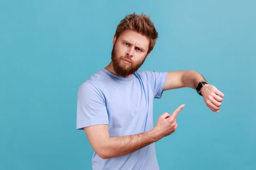 Portrait of anxious nervous bearded man pointing finger on his wristwatch, impatiently checking time, being late, deadline, serious expression. Indoor studio shot isolated on blue background.