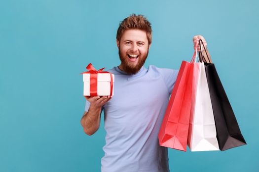 Portrait of smiling satisfied handsome bearded man holding packages and gift box, looking at camera with happy positive expression. Indoor studio shot isolated on blue background.