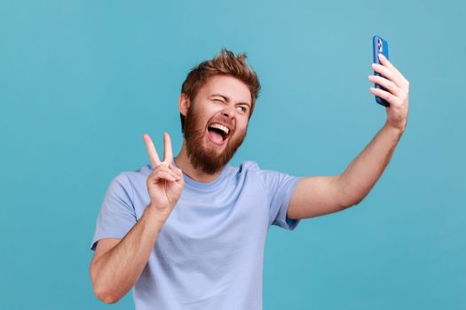 Portrait of successful man taking selfie or talking on video call and showing victory, peace gesture, pleasant online communication via mobile phone app. Indoor studio shot isolated on blue background
