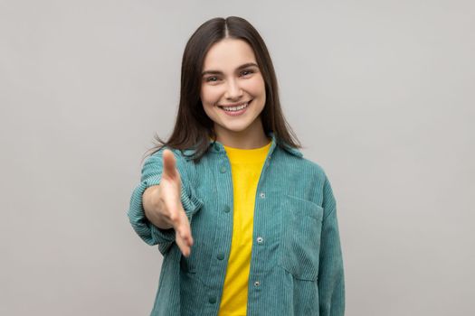 Portrait of happy beautiful young adult woman standing and giving hand to greeting and toothy smile, wearing casual style jacket. Indoor studio shot isolated on gray background.