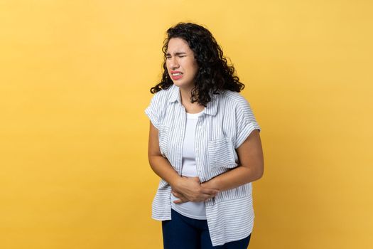 Portrait of unhealthy sick beautiful woman with dark wavy hair standing, holding her belly with hands, stomach cramps or period pain. Indoor studio shot isolated on yellow background.