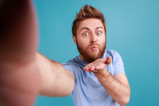 Portrait of romantic flirting bearded handsome man blogger making selfie POV, sending air kisses to followers while having livestream. Indoor studio shot isolated on blue background.
