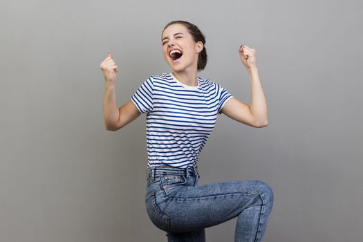 I am champion. Portrait of woman wearing striped T-shirt screaming for joy, raising high her fists, celebrating successful winning, incredible victory. Indoor studio shot isolated on gray background.