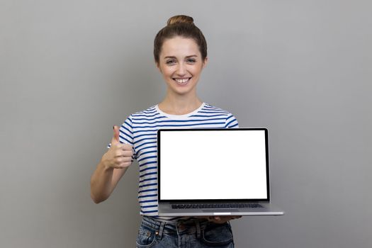 Happy smiling satisfied woman wearing striped T-shirt holding laptop with empty blank display and showing thumbs up, pleased with online application. Indoor studio shot isolated on gray background.