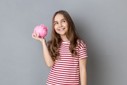 Portrait of adorable pretty smiling little girl wearing striped T-shirt holding pig money box in hands, saving money, banking. Indoor studio shot isolated on gray background.